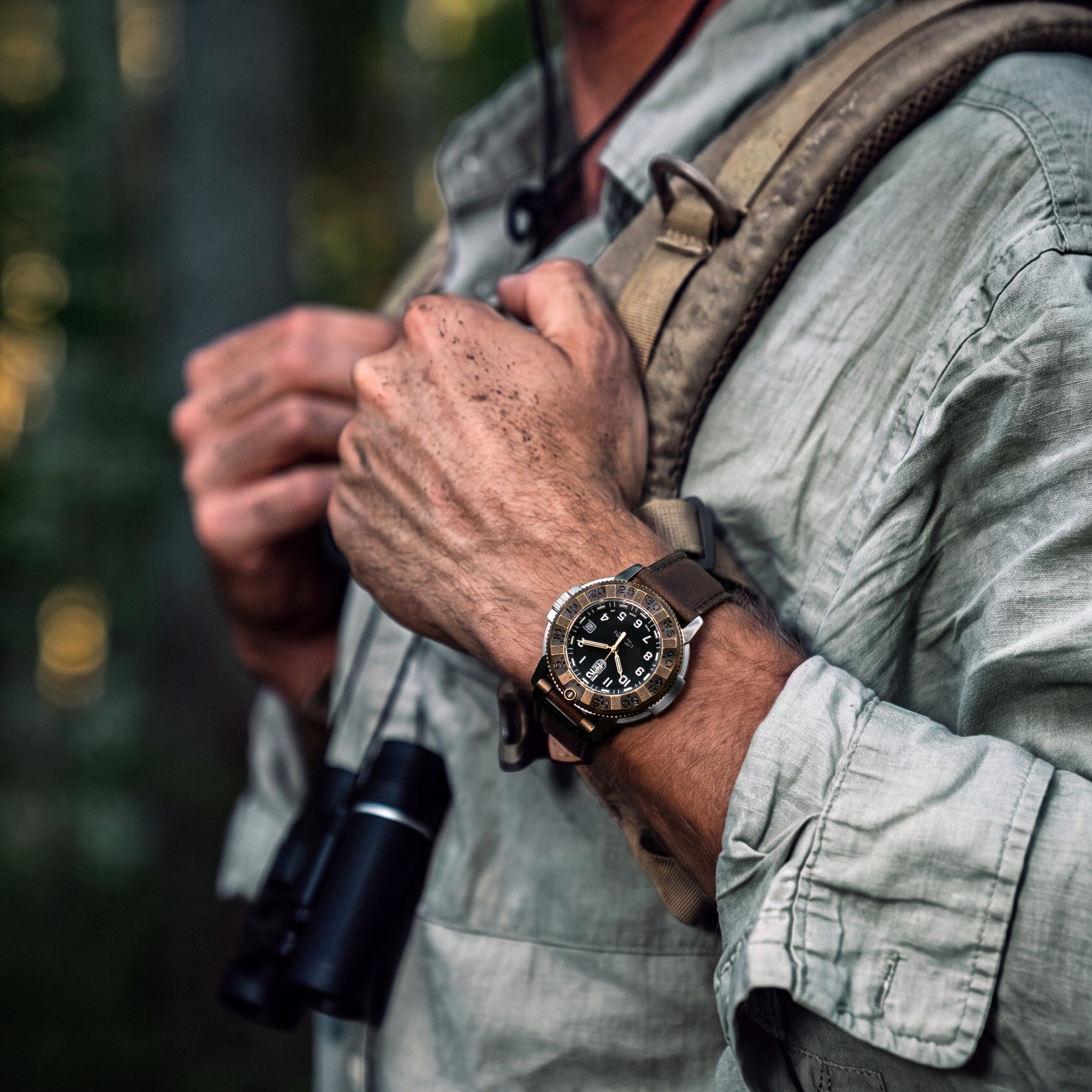 Close-up of an explorer's hands holding his backpack straps, prominently displaying the Luminox Adventure Watch XL.1301.LG on his wrist and binoculars hanging from his neck
