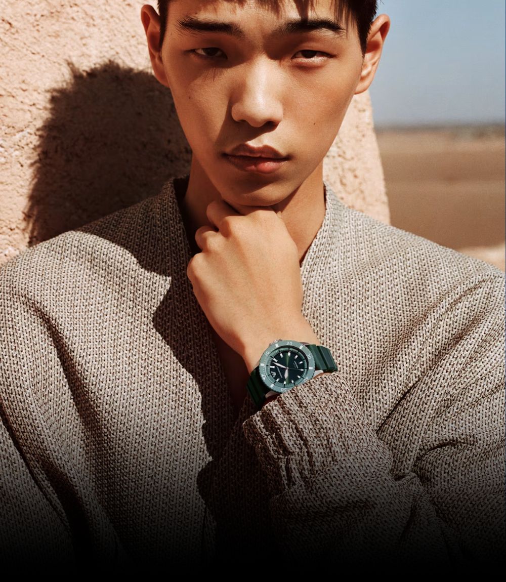 A man wearing a textured gray knit sweater and an Emporio Armani dark green watch with a blue dial, posed against a warm beige stucco wall in a desert-like outdoor setting