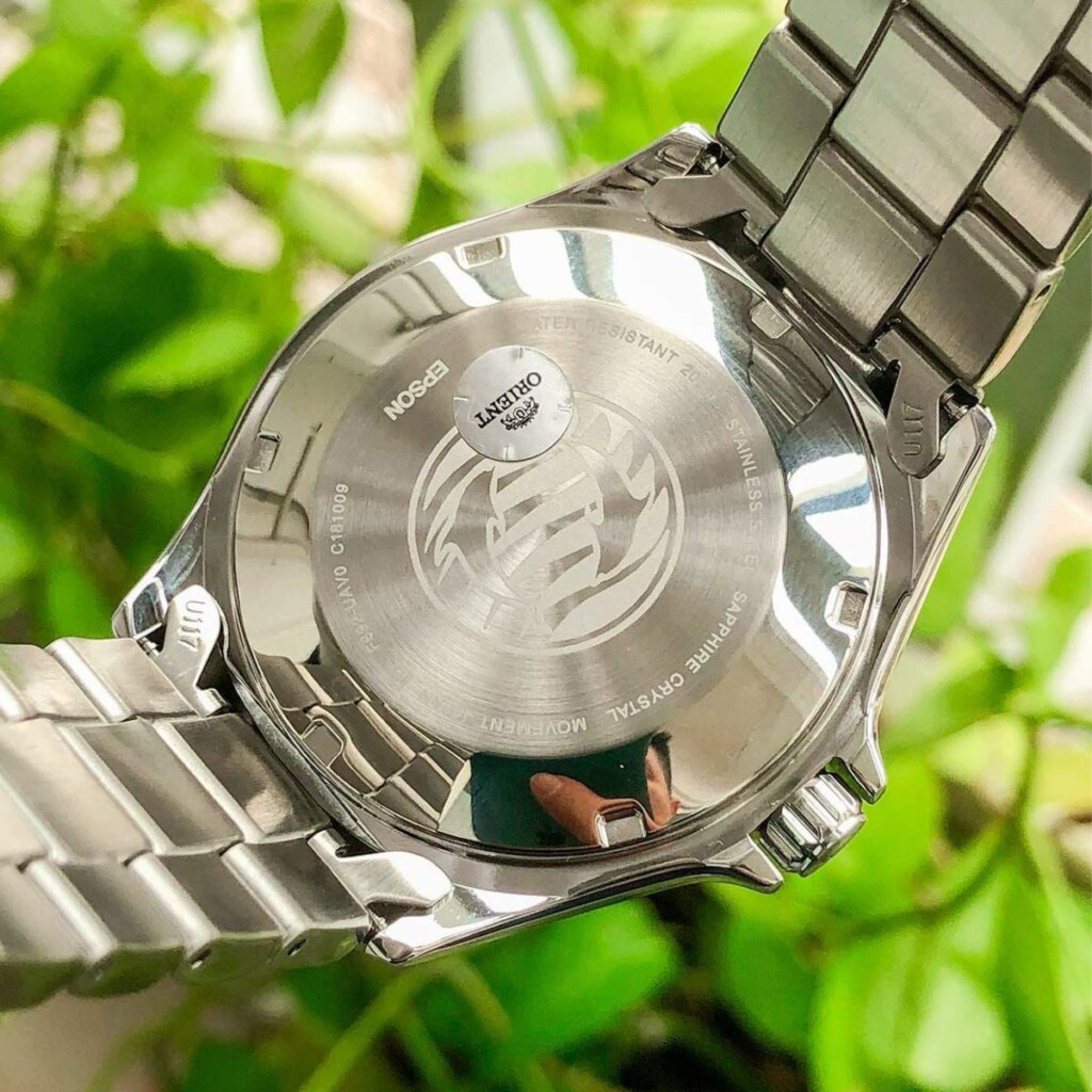 Close-up of a silver watch with brand logo against a blurred green leafy background