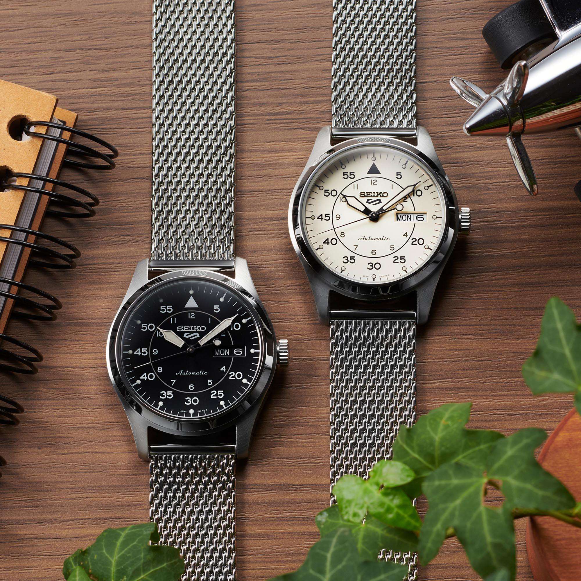 Two Seiko watches with mesh straps on a wooden surface with a plant.