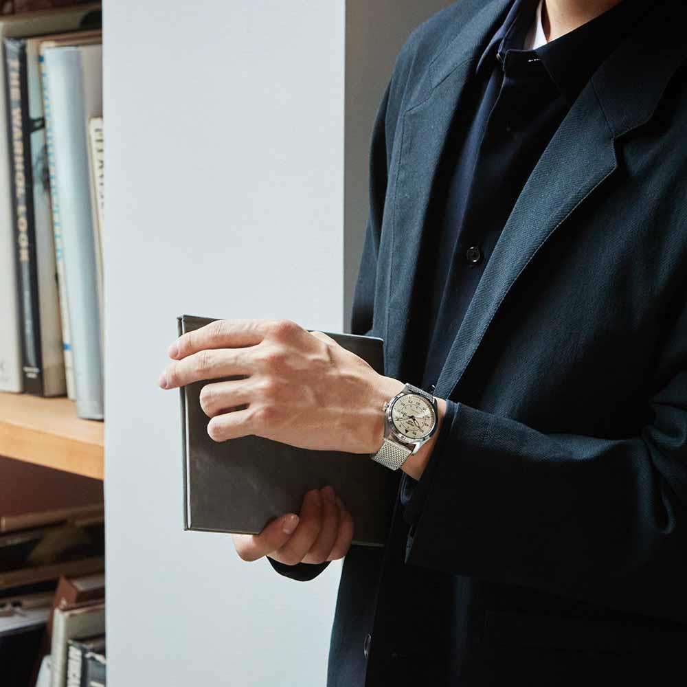 Person holding a book with a watch on their wrist, standing in front of a bookshelf.