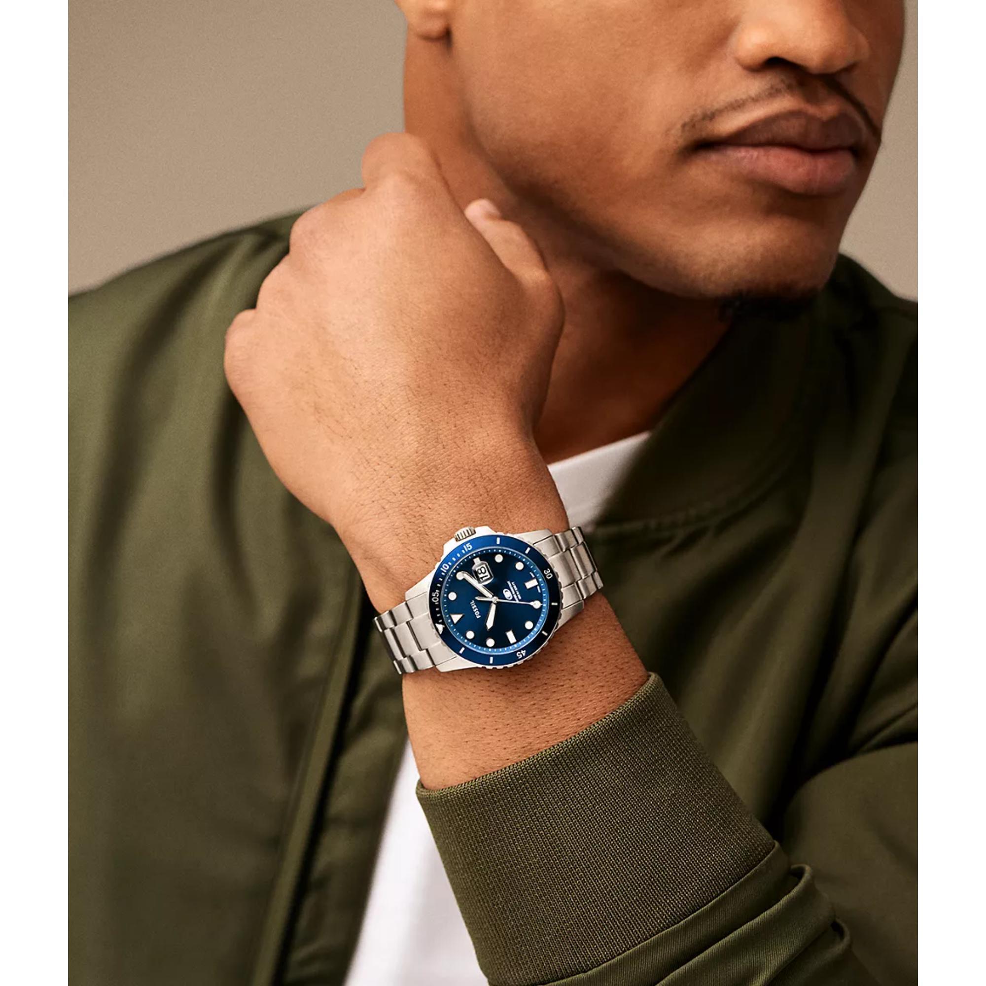 Man wearing a silver watch with a blue face on his wrist, against a neutral background.