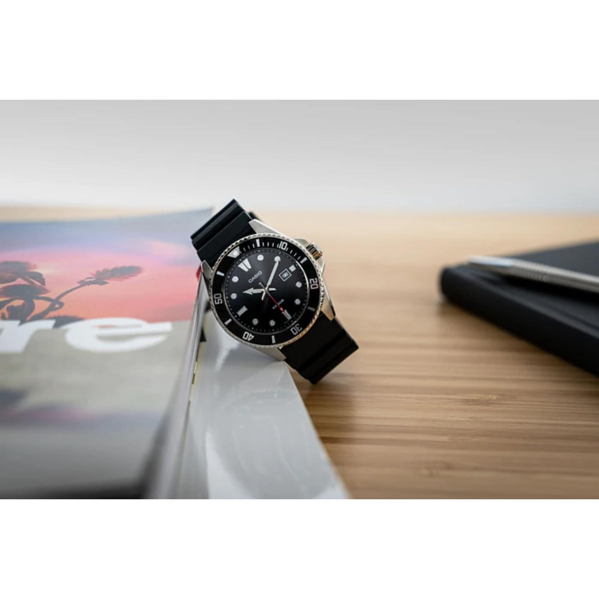Casio Marlin MDV-107-1A1VEF resting against a magazine on a wooden surface next to a notebook and pen