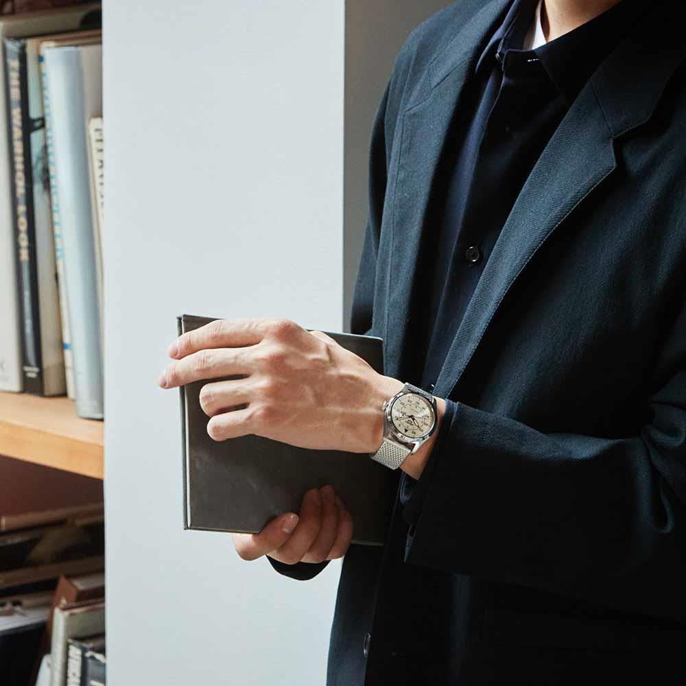 Person holding a book with a watch on their wrist, standing in front of a bookshelf.
