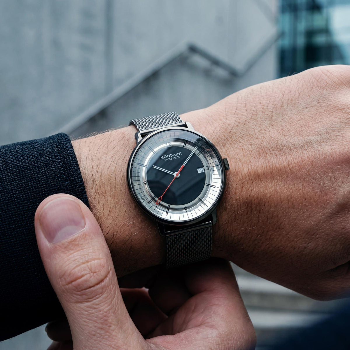 A close-up lifestyle shot of a Mondaine MLE.41280.SM watch on a person's wrist, showing the steel mesh strap and black dial, against a blurred gray concrete background
