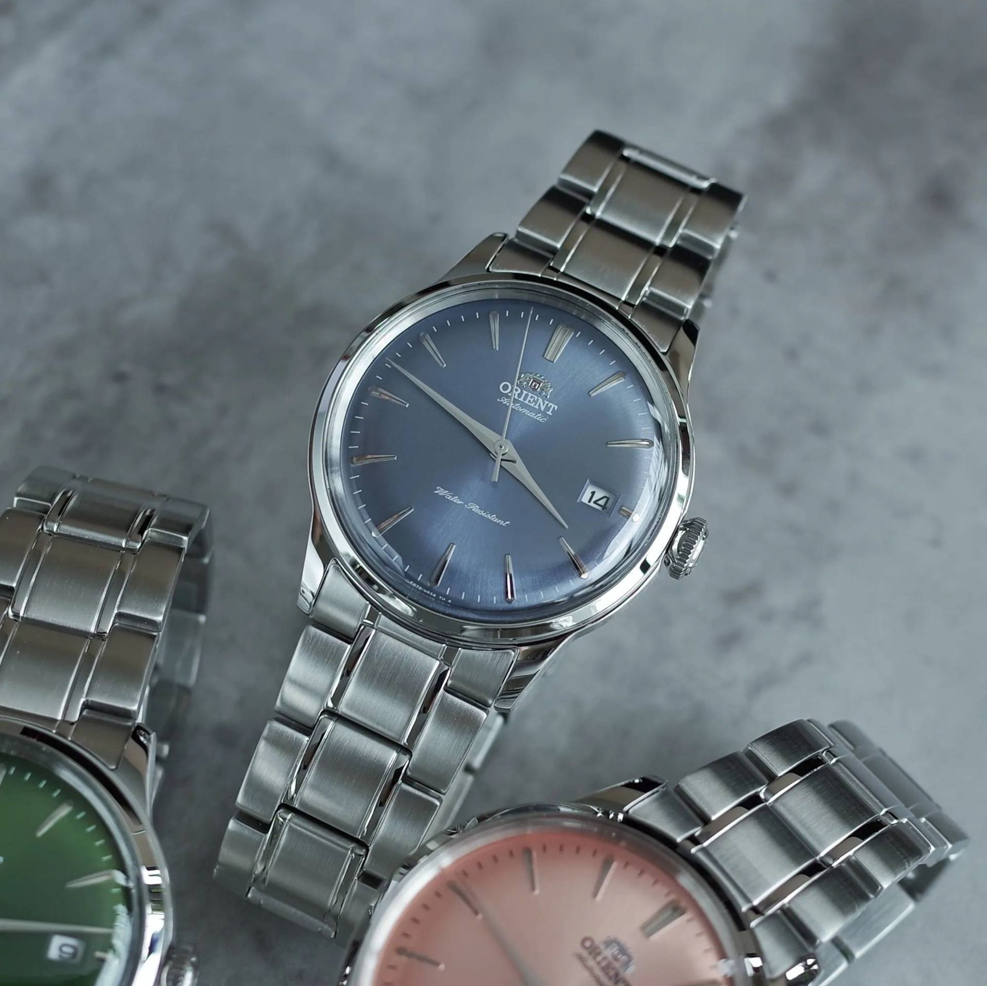 Orient Bambino watch with a blue sunburst dial and steel bracelet, pictured next to green and pink versions.