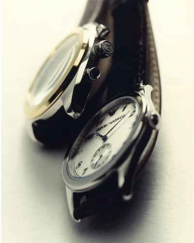 Two Emporio Armani watches with black leather straps, one featuring a gold-tone case and bezel, and the other a silver-tone case, displayed diagonally on a light gray background