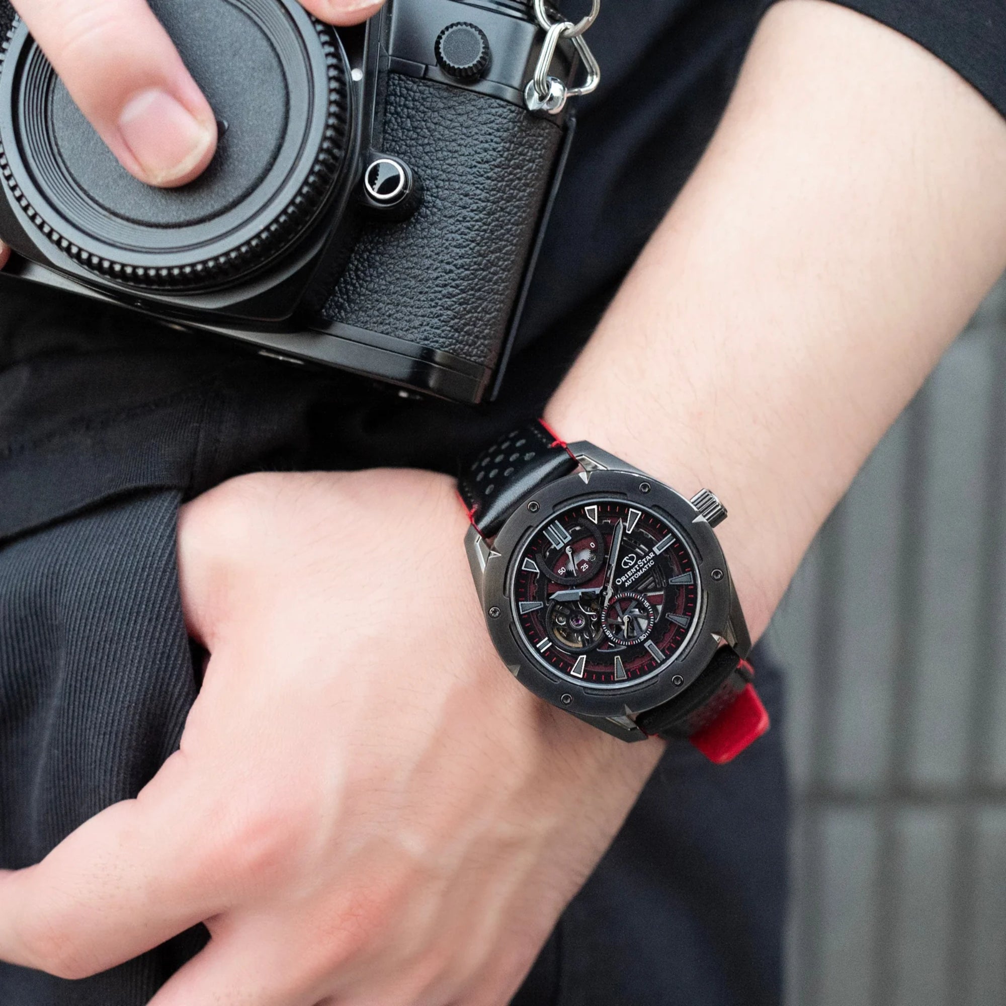 Orient Star RE-AV0A03B00B skeleton watch worn on wrist with black camera in lifestyle shot