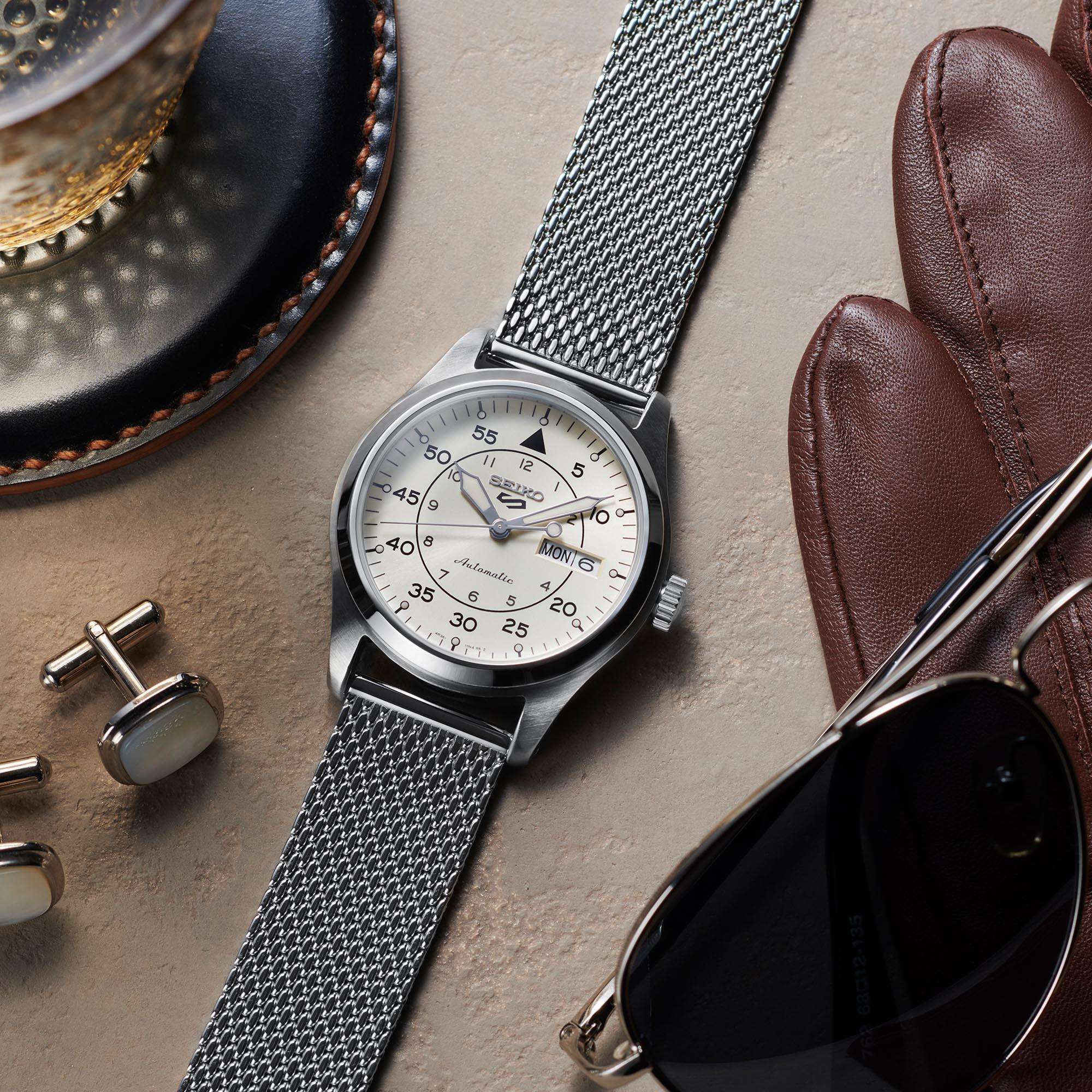 Silver watch with mesh strap on a textured surface with sunglasses, gloves, and cufflinks.