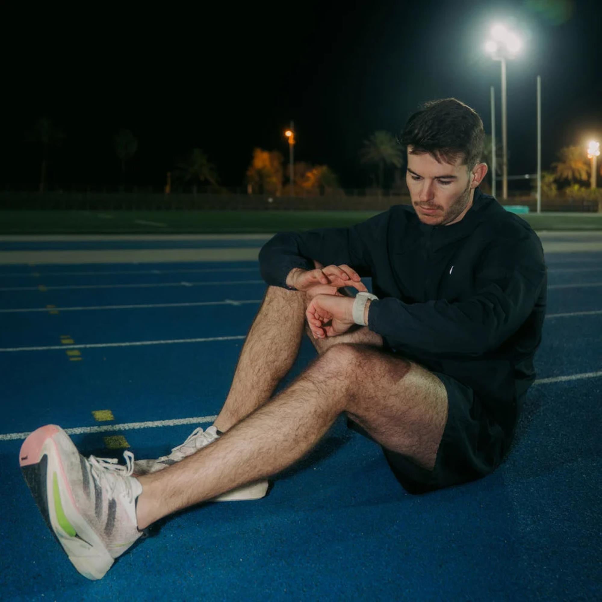 An athlete sitting on a running track at night checking a Suunto watch with a Frost Gray textile strap lifestyle view