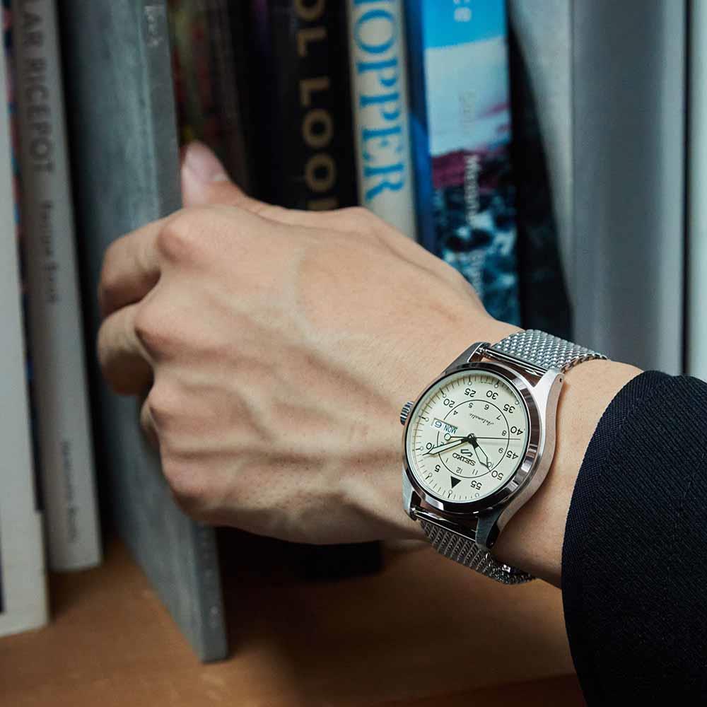 Hand wearing a silver watch with a detailed face, against a background of books.