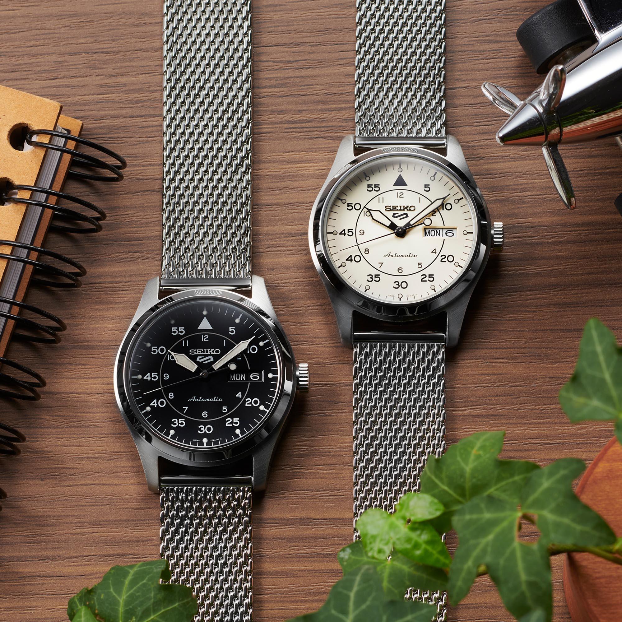 Two Seiko watches with mesh straps on a wooden surface with a plant.