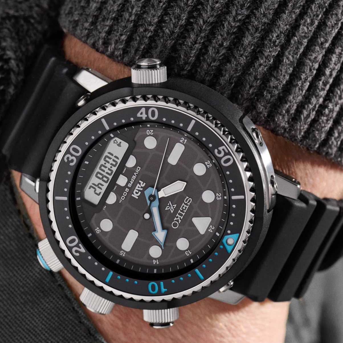 Person wearing Seiko Prospex Solar ‘Arnie’ Hybrid PADI SNJ035P1F wristwatch with a black face and blue accents on a blurred background